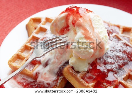 freshly baked Belgian waffles with ice cream and strawberry jam in a white plate with a metal fork on a red table