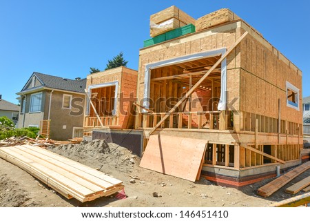 A Single Family Home Under Construction. The House Has Been Framed And Covered In Plywood. Stacks Of Board Timber In Front And Stack Of 2x4 Boards On The Top.