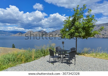 Dining table and chairs on picnic area with panoramic view at Okanagan lake and lonely tree by side.
