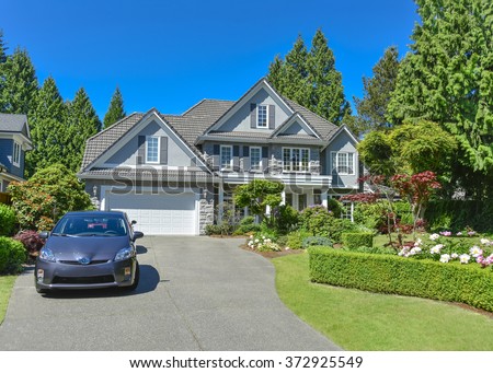 Luxury residential house with green hedge and landscaping in front. Family house surrounded by trees with blue sky background. Suburban house with double garage and car parked on concrete driveway.