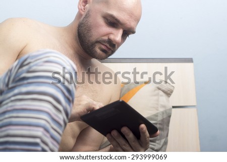 Young handsome bald guy, without shirt, using at tablet in bed.