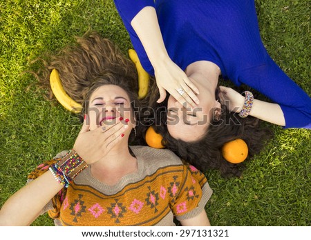 Above view of smiling happy girls, laying on a grass. Making fun with bananas, and oranges.