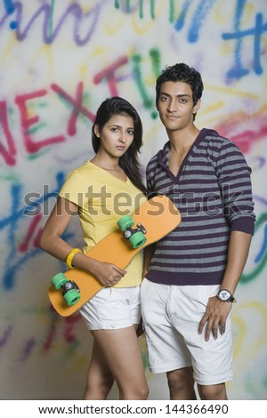 Portrait of a couple standing with a skateboard in front of a graffiti covered wall