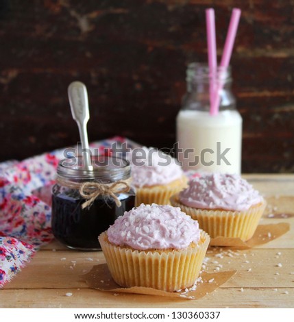 Three cupcakes with blackcurrant jam and coconut and cream cheese frosting in paper linens and a jar of jam and milk bottle with straws on a wooden surface