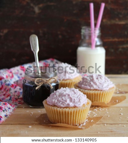 Three cupcakes with blackcurrant jam and coconut and cream cheese frosting in paper linens and a jar of jam and milk bottle with straws on a wooden surface