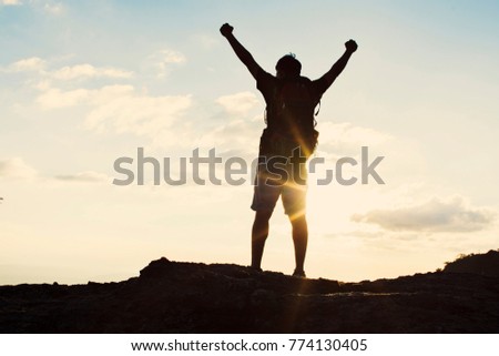 Man with backpack putting his hands up  and standing on cliff at sunset time, sun rays on background with clouds, warm tone and almost in silhouette photo