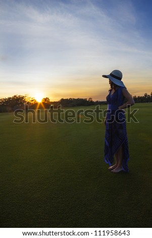 A woman with a blue dress and sun hat standing on a green field of grass watching the sun set. Sun has a flair and sky has wispy clouds.