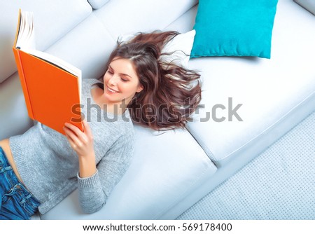 Girl reads a book lying at white soft sofa. Culture lifestyle. Leisure. Top view. Young woman reading art literature.