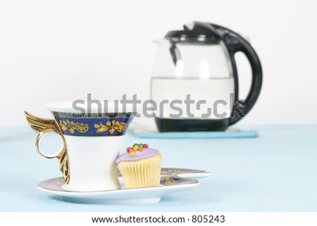 A cup of coffee or tea and a mini cupcake.    Kettle in background out of focus.