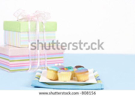 Miniature cakes sitting on a white plate on a blue tablecloth.  Pastel boxes tied with pale pink satin and gauze ribbon in background.  Focus is to the foreground and cakes only.  Space for a message.