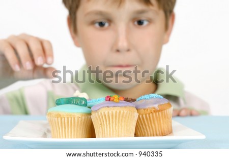Mmmmm - photo shows a boy eyeing off some iced cup cakes.  He has his hand outstretched to take one.    The focus is to the cakes.