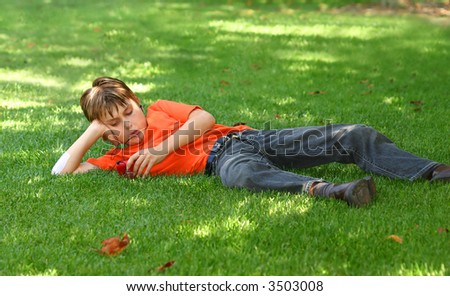 a young boy sprawled out on the shaded grass in the park, listens to music on an mp3 music player.