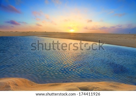 The summer sun rises over golden sand dunes and azure blue waters.  This is the tide flowing in quite rapidly through a narrow channel
