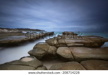 Weathered rocks, ocean channel and rock craters at Soldiers Beach Australia under a stormy dark sky.  Norah Head lighthouse in distance.