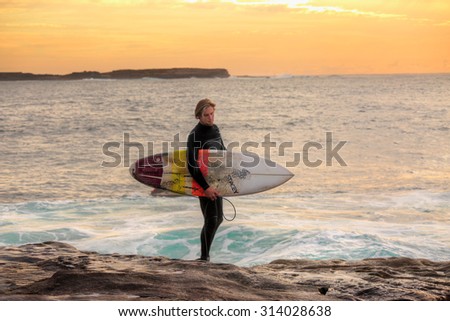 CAPE SOLANDER, AUSTRALIA - 27 AUGUST, 2015; Surfer at sunrise carrying a Stacey Roach surfboard and wearing an O\'Neill wetsuit, standing on rocks at  Cape Solander.