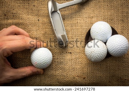 A hand holding a golf ball near a putter and three other golf balls