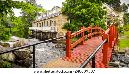 Stream runs past a mill and under a red foot bridge.