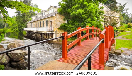 Stream runs past a mill and under a red foot bridge.