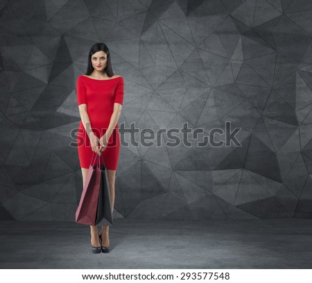 Beautiful brunette woman in a red dress is holding fancy shopping bags. Contemporary background.