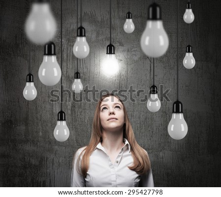 Charming lady is looking upward at the hanging light bulbs. A concept of searching new ideas. Dark concrete background.