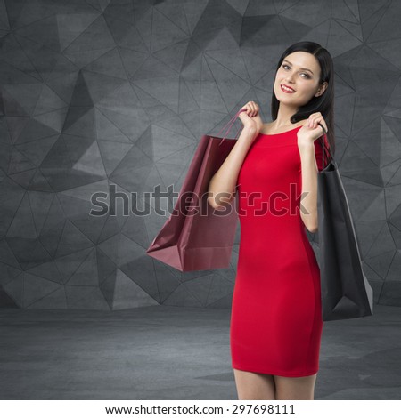 Beautiful brunette woman in a red dress is holding fancy shopping bags. Contemporary background.