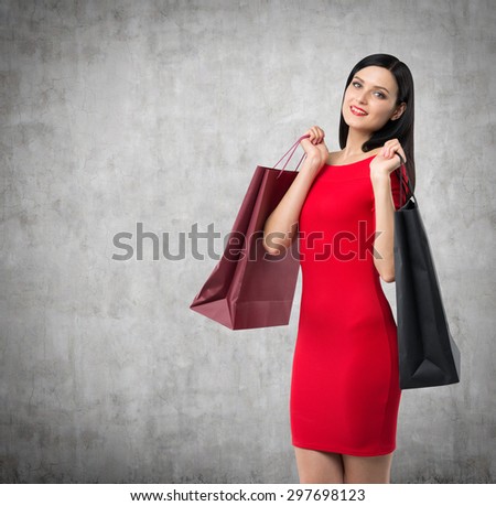 Beautiful brunette woman in a red dress is holding fancy shopping bags. Concrete background.