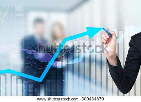 A hand is drawing a growing arrow on the glass scree, Blue dark background with financial graphs. Business couple in blur on the background.