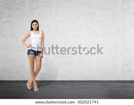 Full length portrait of a brunette woman who is in a white tank top and blue denim shorts. Room with the concrete wall.