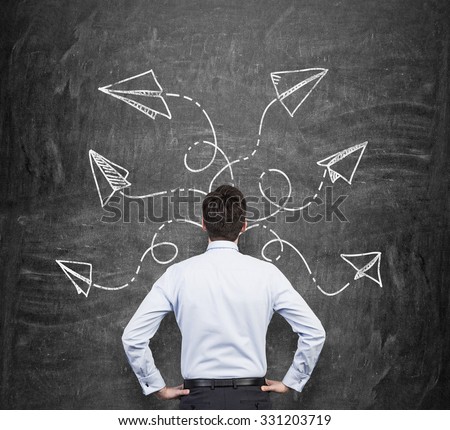 Rear view of a man in formal clothes who is pondering about possible solutions of the complicated problem. Many arrows with different directions are drawn around his head on the black chalkboard.