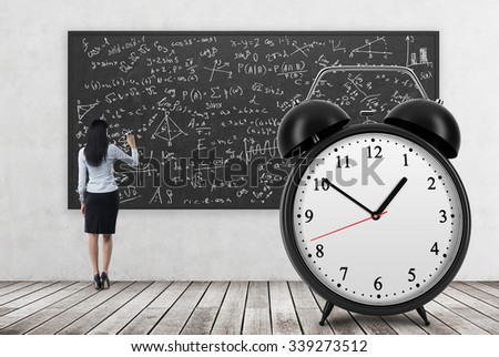 Rear view of business lady who is writing math formulas on the black chalkboard. The huge alarm clock is on the foreground. A concept of time and quantitative science. Wooden floor and concrete wall.