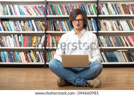 smiling young man in white shirt sitting on the floor with crossed legs in the library with a laptop on his knees, looking at the screen, book shelves at the background, a concept of studying.