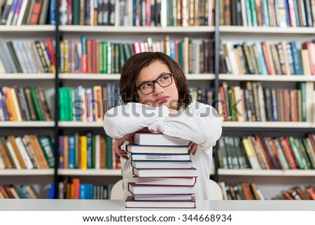 young man sitting at a desk in the library with his arms on a big pile of books and chin on them, head half turned to the top,  thinking,  a concept of knowledge, blurred books at the back