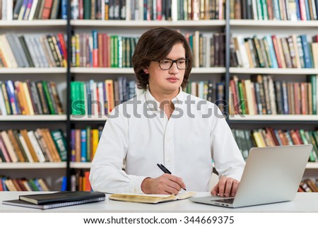 serious young man with dark hair  sitting at a desk in the library making notes, laptop and organiser on the table, looking at the screen, a concept of studying, blurred books at the back