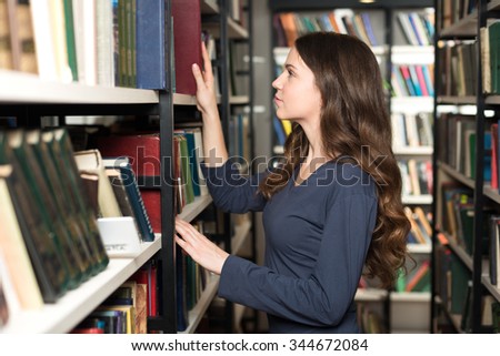 young lady with loose long dark hair choosing a book between shelves in the library, touching a book, side view, a concept of studying and choice