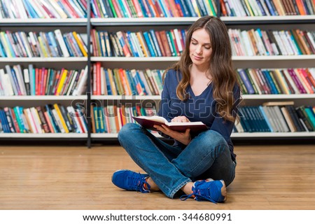 smiling young girl  sitting on the floor in the library reading an open book on her knees, left-centered, blurred bookshelves at the background, a concept of reading