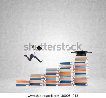 Young man with a folder in hand running up stairs made of piles of books of different size, graduation hat on the highest, concrete background. Concept of higher education.