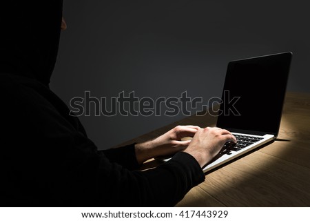 Man with hood on sitting at wooden desk and hacking notebook with blank screen. Mock up