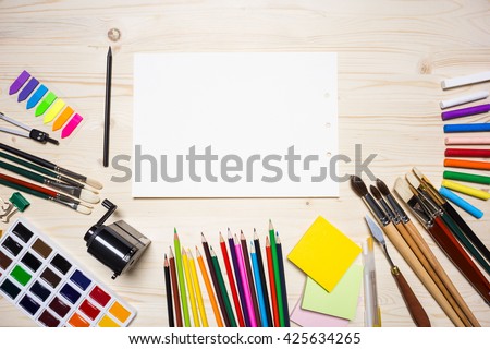 Top view of light wooden desktop with piece of white paper and various colorful drawing tools. Mock up