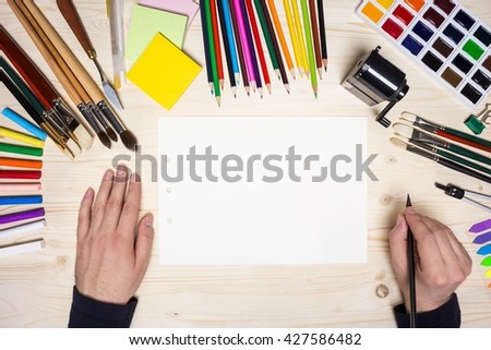 Top view of artist\'s hands with pencil on wooden desktop with blank paper and drawing tools. Mock up