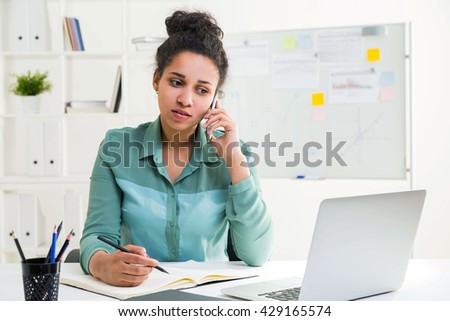 Pretty african american woman having phone conversation at office desk with laptop, notepad and other stationery items