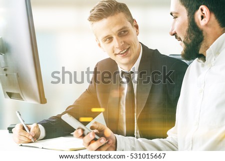 Portrait of two business partners sitting at a table together and working. One is holding his smartphone. The second is writing but can\'t draw his eyes from the colleague. Toned image