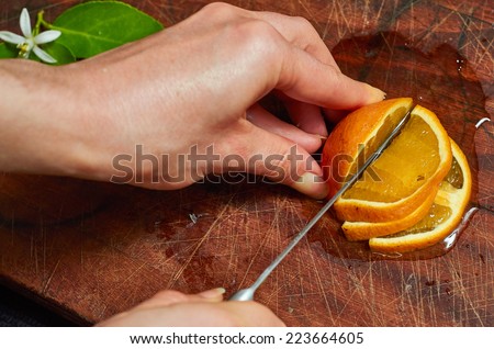 A quarter of an orange on a wooden board being sliced with a knife being held by a woman\'s hands.