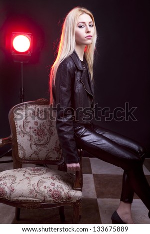 Girl in leather jacket with a red light behind sitting next to a vintage chair studio shot