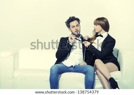 Young boy sitting on the white sofa with a cigar in his mouth while his girlfriend pulls him by the tie