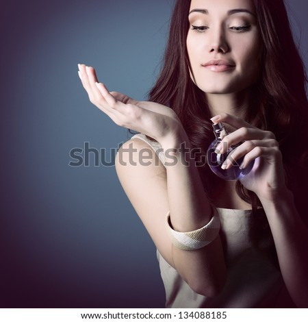 Woman with perfume, young beautiful girl holding bottle of perfume and smelling aroma, over blue purple background