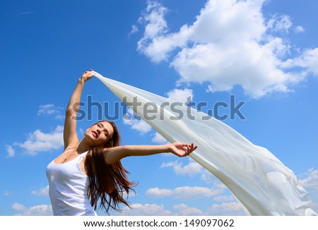 Happy young woman holding white scarf with opened arms expressing freedom, outdoor shot against blue sky