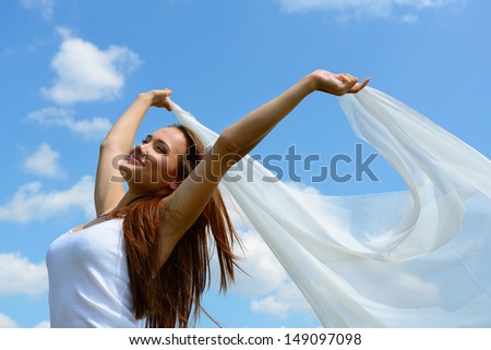 Happy young woman holding white scarf with opened arms expressing freedom, outdoor shot against blue sky