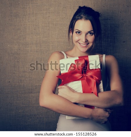 Portrait Of Attractive Cheerful Girl In Sleeveless Sports White Shirt Holding Gift Box With Red Bow Over Canvas Background, Toned And Noise Added