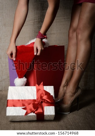 Beautiful young woman with gift box, santa\'s hat and colored shopping bags, long legs on high heels closeup over canvas background, glamour holiday shopping concept, image toned