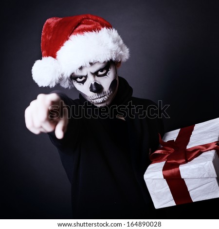 Holiday background of halloween person with terrible skull make-up in santa\'s hat with gift box pointing at you over black background